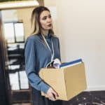 Professional woman carrying a cardboard box filled with documents and a small lamp, walking through an office hallway.