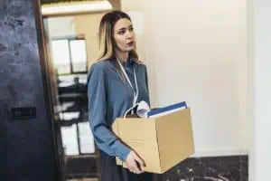 Professional woman carrying a cardboard box filled with documents and a small lamp, walking through an office hallway.