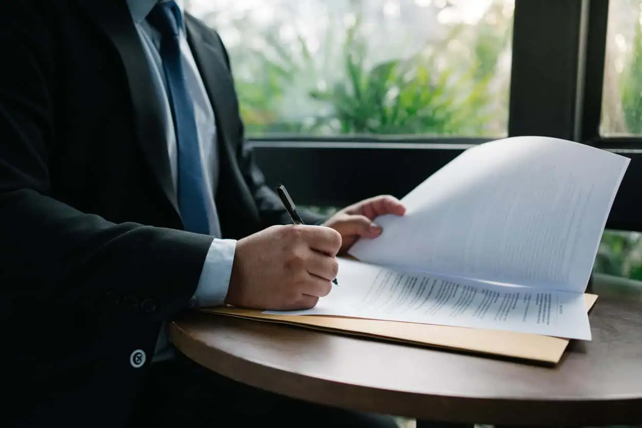 Person in a dark suit signs documents with a pen at a round wooden table.