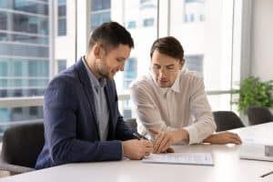 Two businessmen sign a document at a bright office table with a laptop nearby.